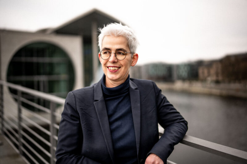 Ulle Schauws lehnt an einem Geländer vor einem Fluss und trägt einen dunklen Blazer und ein dunkles Oberteil