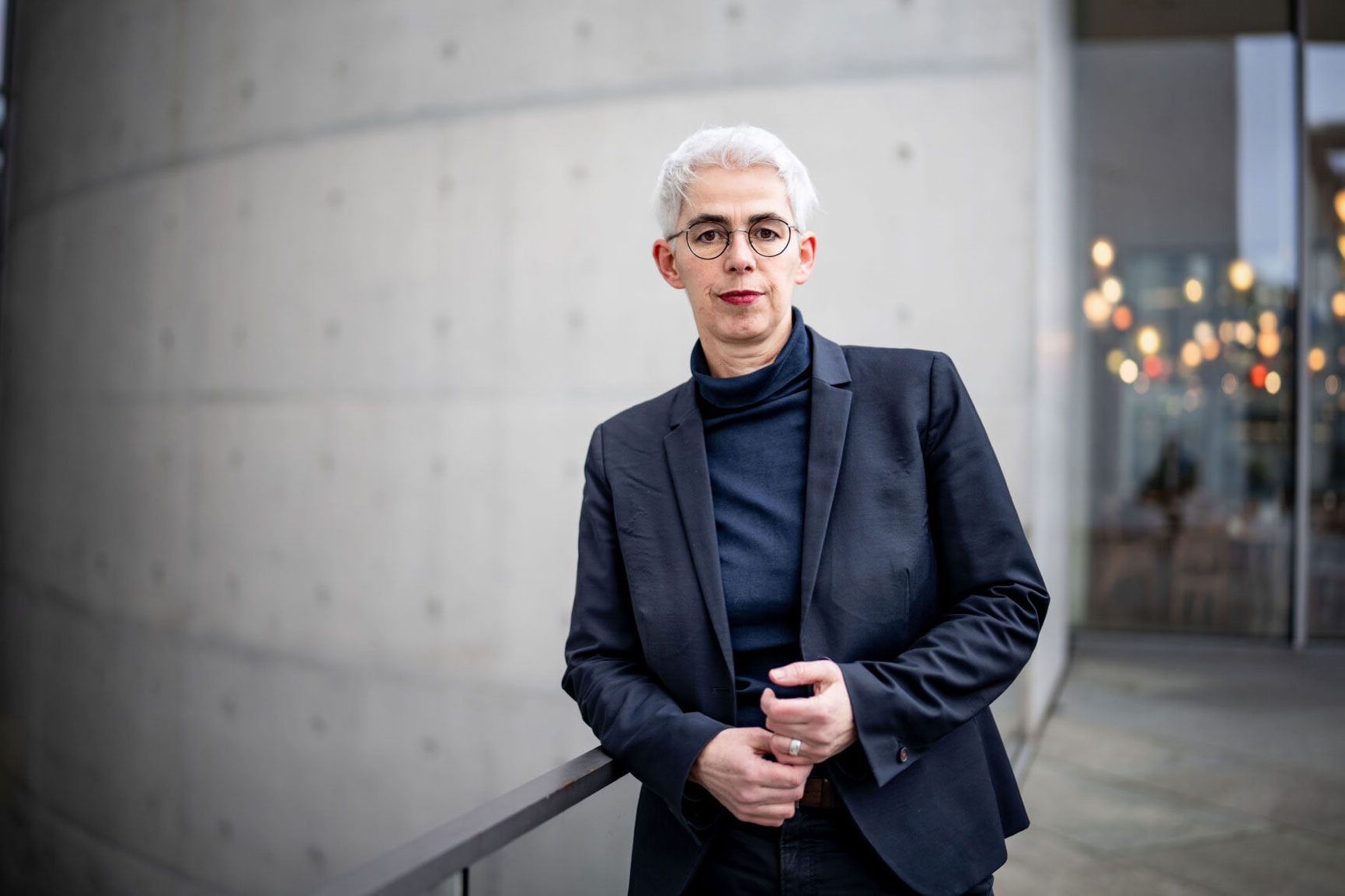 Ulle Schauws lehnt an einem Geländer vor einer Betonwand und trägt einen dunklen Anzug mit Rollkragenpullover