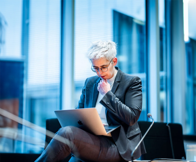 Ulle Schauws sitzt in einem Anzug auf einem Stuhl und arbeitet an einem Laptop in einem modernen Bürogebäude