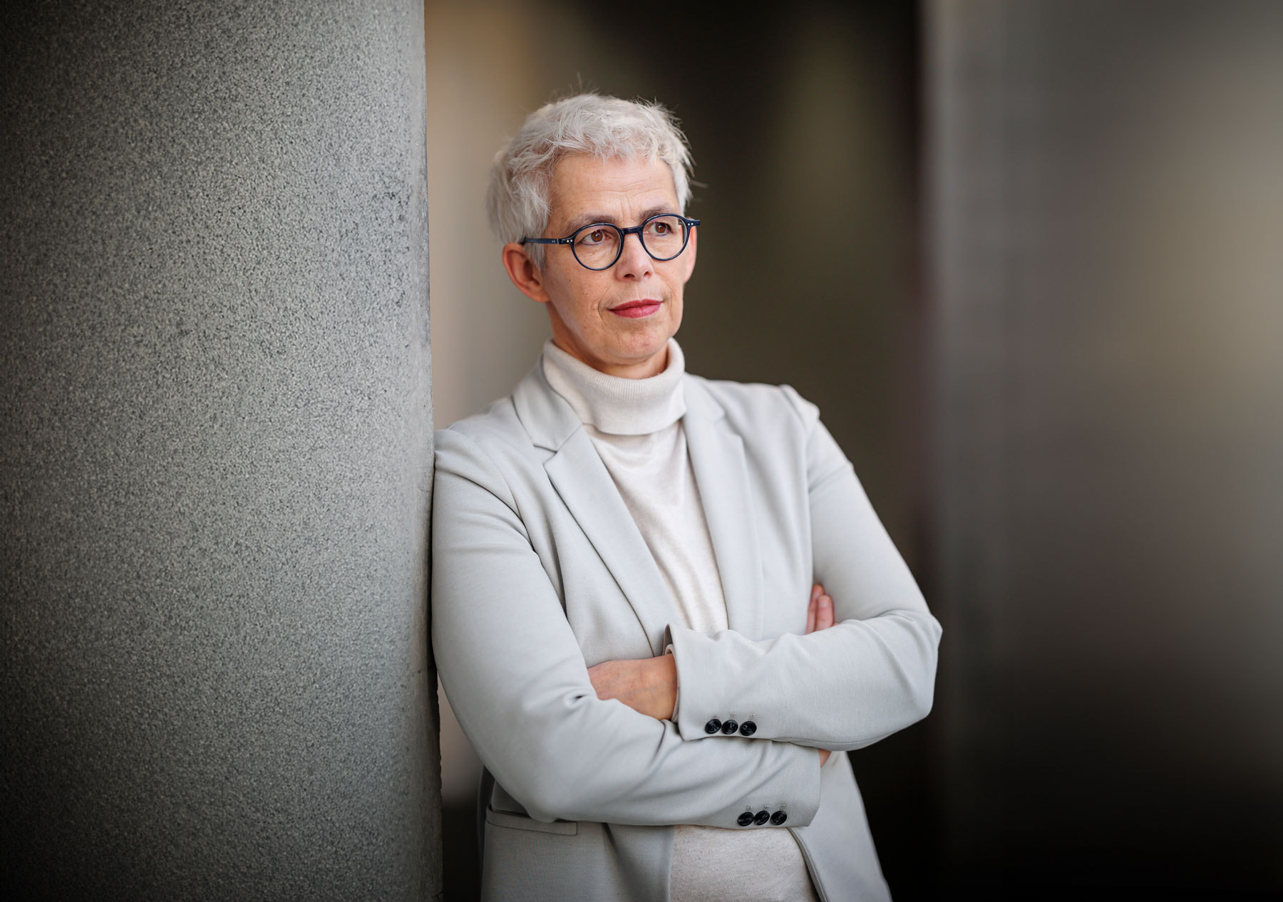 Ulle Schauws lehnt mit verschränkten Armen an einer Wand und trägt einen hellen Blazer über einem Rollkragenpullover