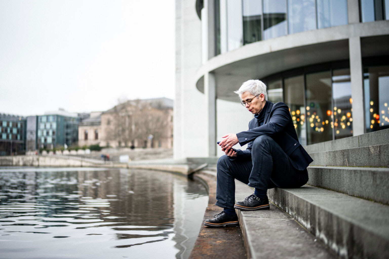 Ulle Schauws sitzt auf einer Treppe neben einem Gewässer vor einem modernen Gebäude mit großen Glasfenstern