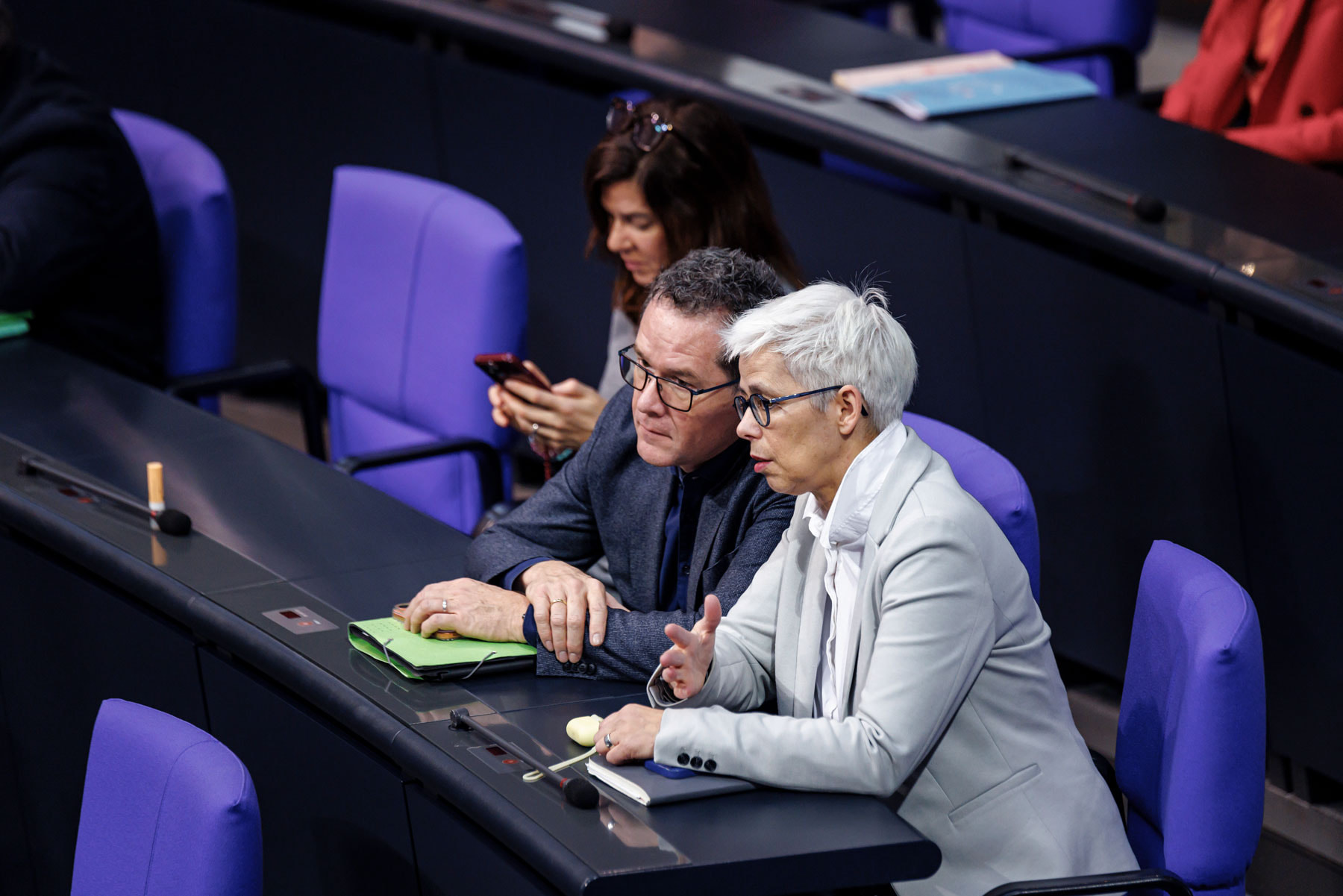 Ulle Schauws und zwei andere Grünenpolitiker sitzen in einem Sitzungssaal mit blauen Sitzen, zwei von ihnen unterhalten sich, eine nutzt ein Smartphone